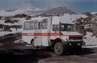 Weißer Geländebus mit rotem Streifen parkt for schneebedeckter Vulkanlandschaft. Tür geöffnet, im Hintergrund sind Menschen.